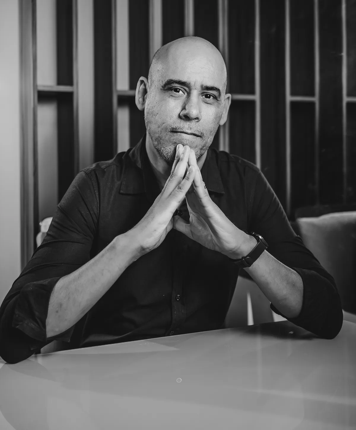 Fábio Pedroso, de camisa preta, sentado à mesa com as mãos unidas próximas ao rosto.