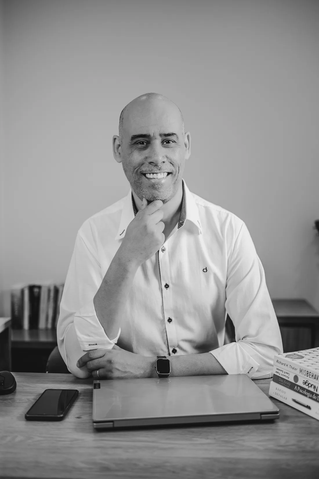 Fábio Pedroso de camisa branca, sorrindo, sentado à mesa com uma das mãos apoiada no queixo.